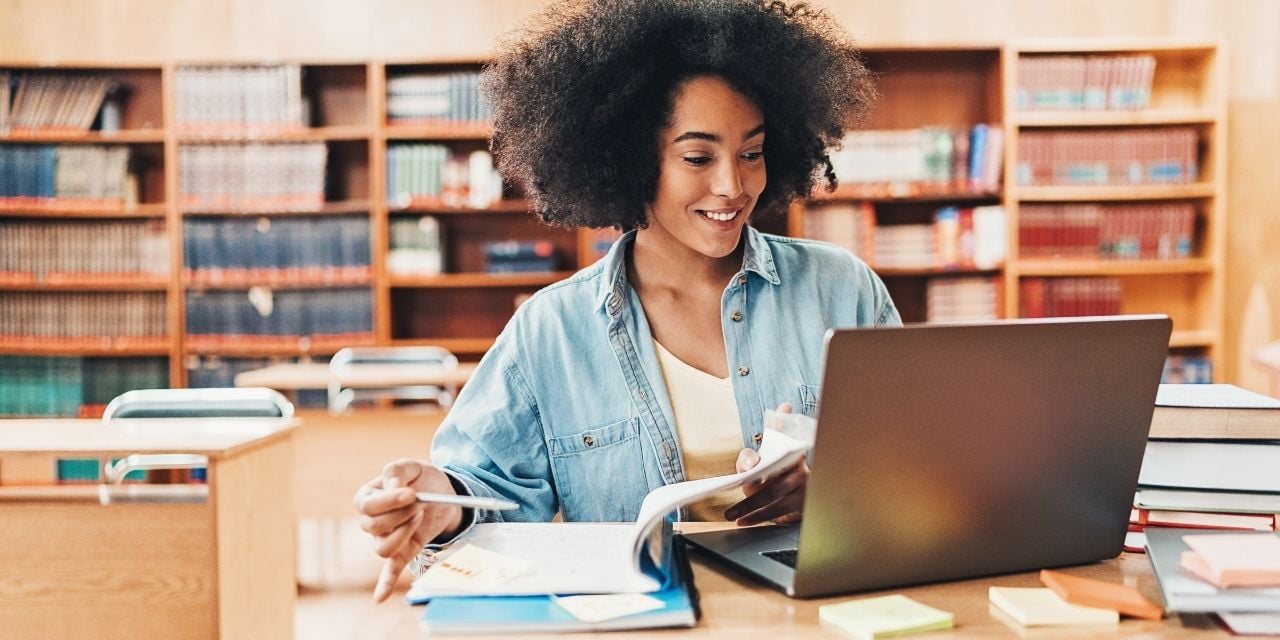 vrouw die met een boek en laptop zit in een bibliotheek